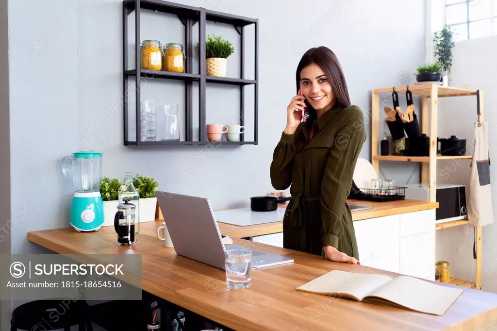 Businesswoman talking on smart phone while standing in kitchen at home