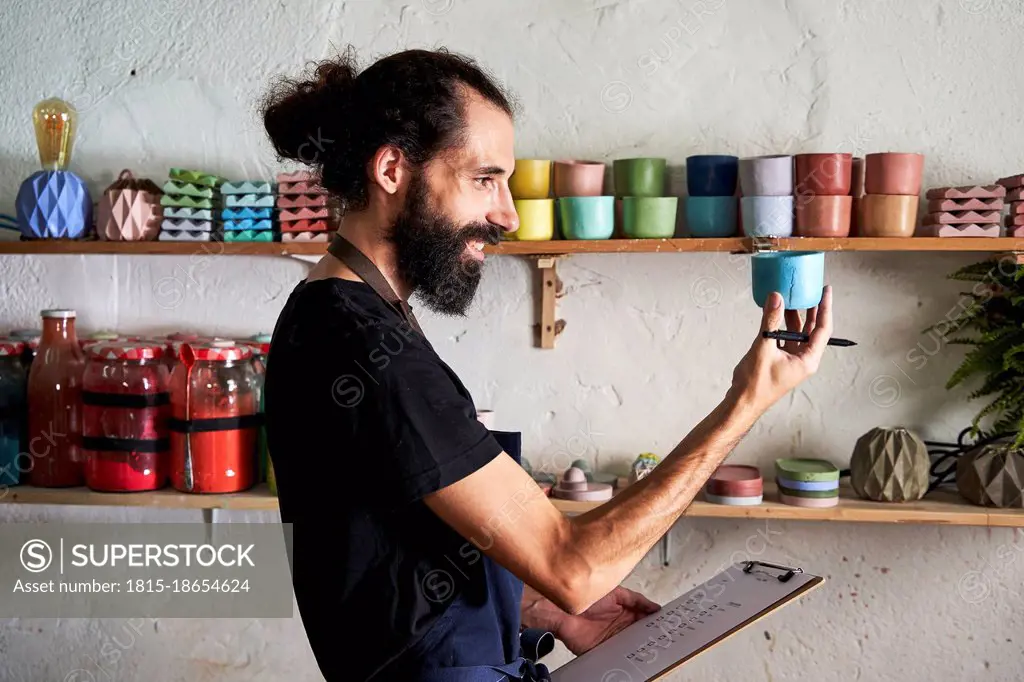 Smiling male entrepreneur holding clipboard while examining concrete container in workshop