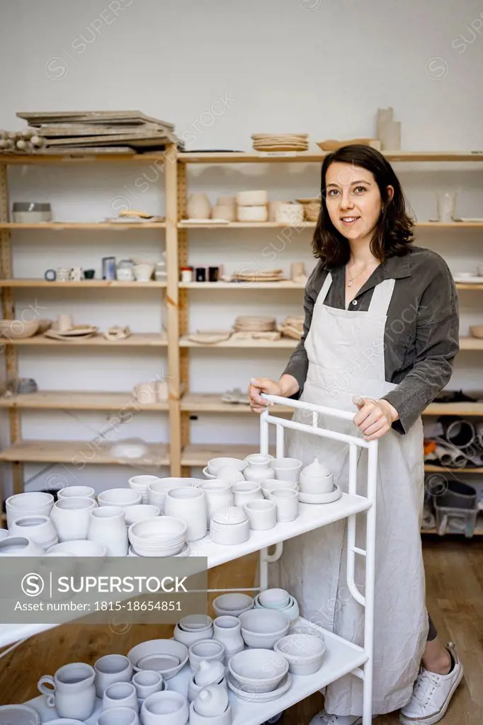 Smiling young craftswoman with pottery cart in workshop