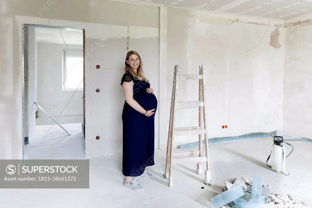 Smiling pregnant woman touching abdomen at house under renovation