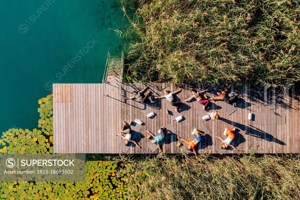 Men and women practicing yoga on jetty by lake during sunny day