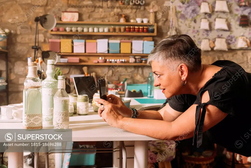 Mature woman photographing craft product through smart phone at workbench