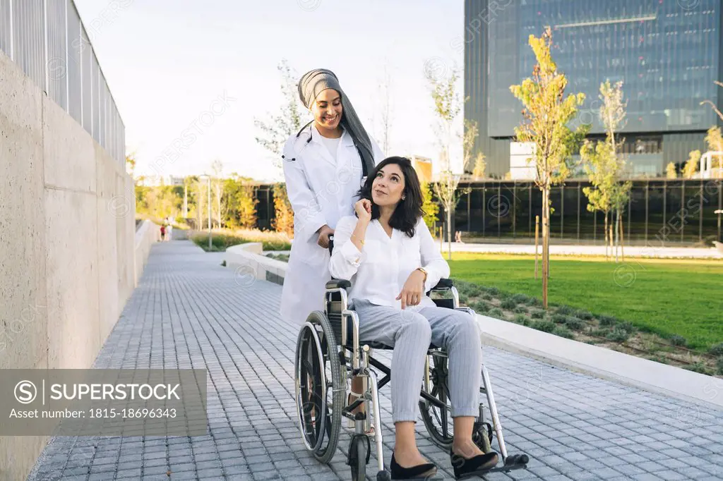 Disabled woman talking with female doctor pushing wheelchair on footpath