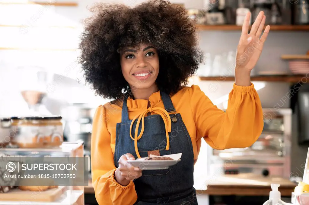 Smiling waitress holding pastry in plate gesturing at cafe