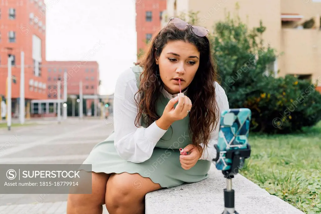 Young curvy influencer recording on smart phone while applying lipstick