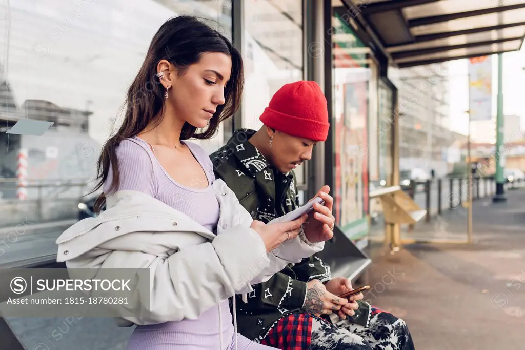 Couple using smart phones sitting on bench at bus stop