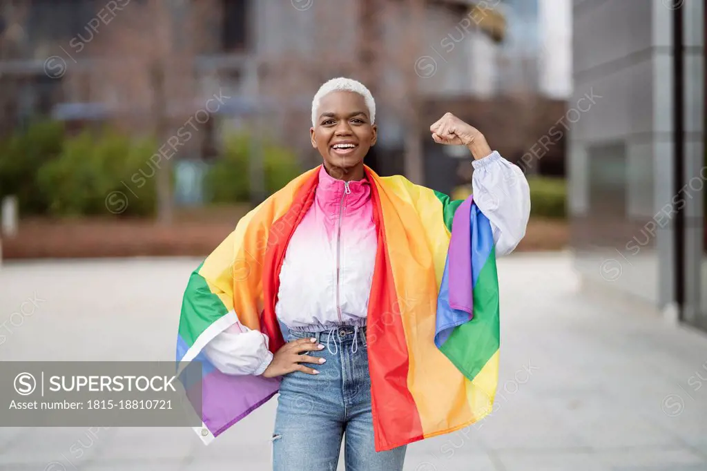 Young woman wrapped in rainbow flag flexing muscle with hand on hip