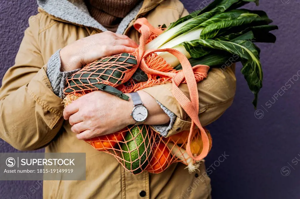 Woman holding mesh bag with vegetables in front of purple wall
