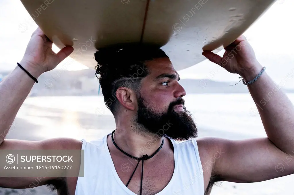 Bearded man carrying surfboard on head at beach