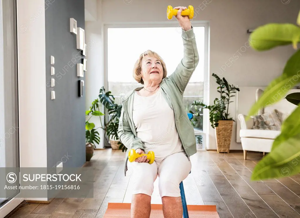 Senior woman with hand raised holding dumbbell sitting on chair at home