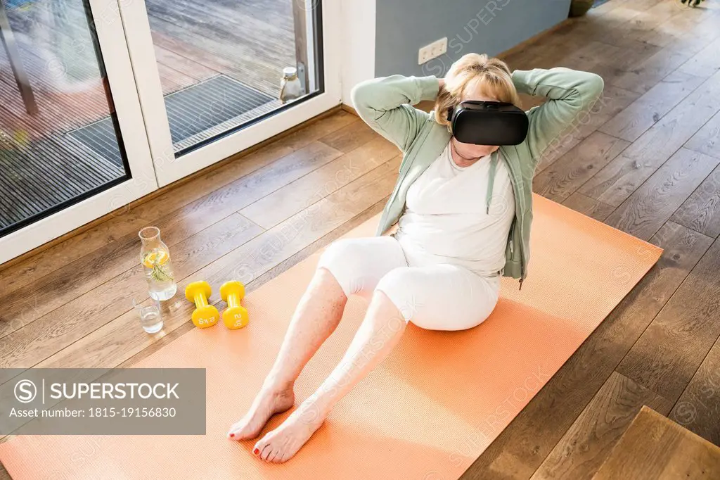 Senior woman wearing virtual reality simulator exercising with hands behind head at home