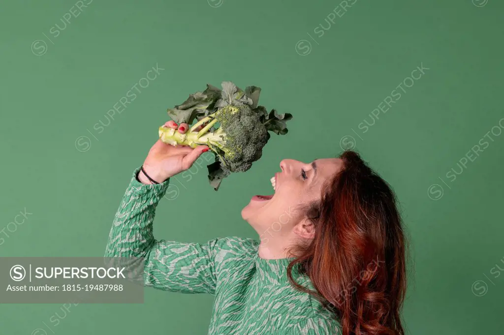 Redhead woman with mouth open holding broccoli standing against green background