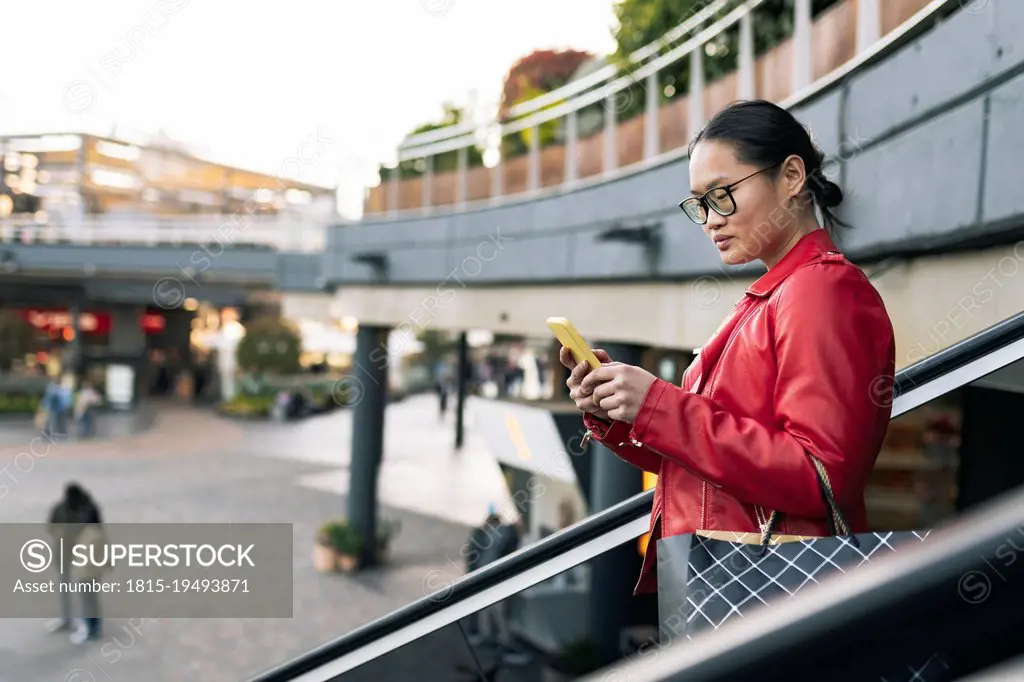 Young woman with shopping bag using smart phone moving down on escalator