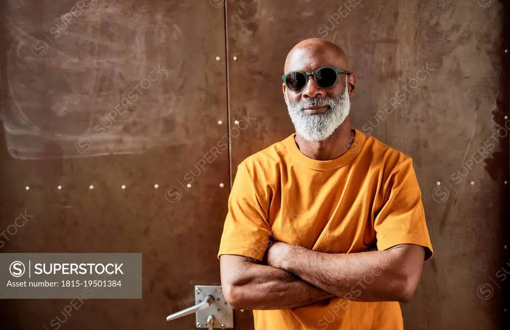 Bald man wearing sunglasses standing with arms crossed in front of door -  SuperStock