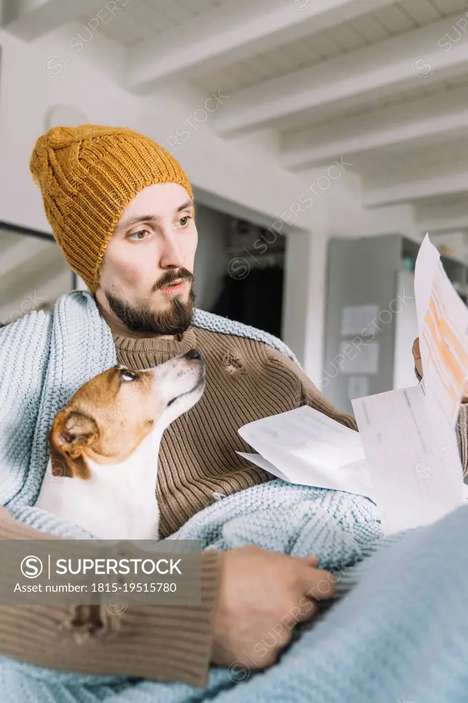 Wickede, NRW, Deutschland. Ein junger Mann trÇÏgt eine StrickmÇ¬tze Zuhause mit einem Hund Jack Russell Terrier schaut erstaunlich auf eine Rechnung a...