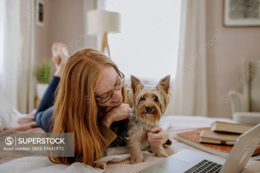 Smiling woman playing with pet dog lying on bed at home