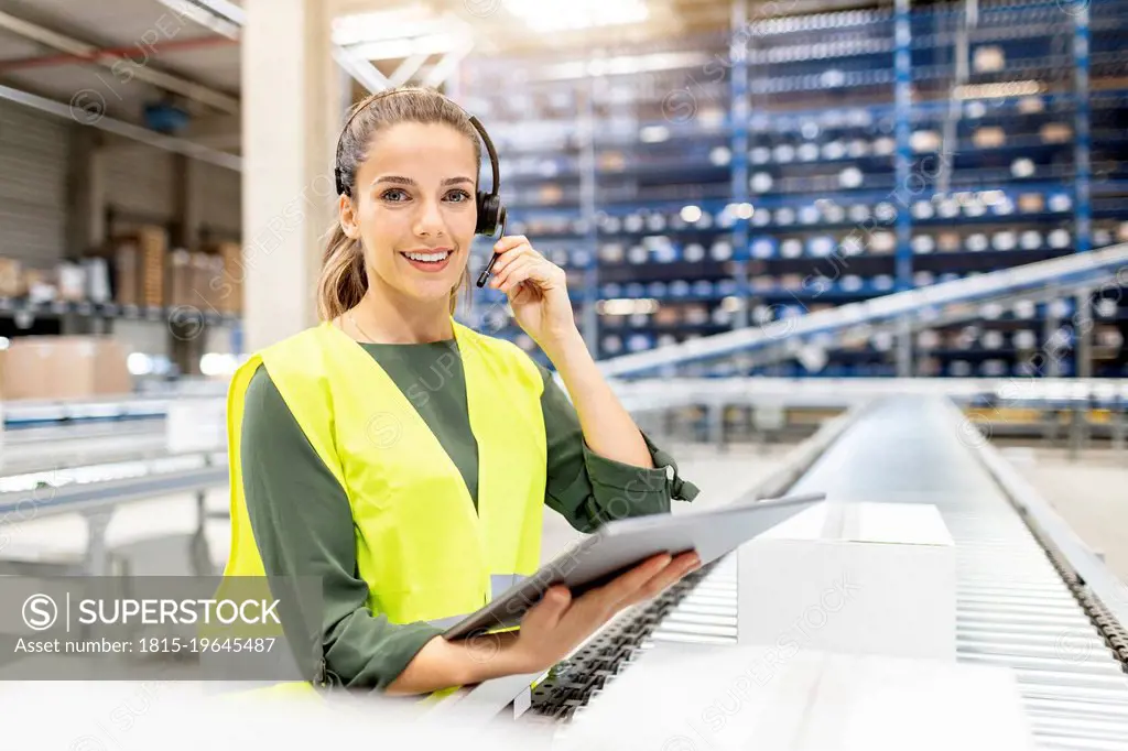 Smiling worker wearing headset standing with tablet PC by conveyor belt in warehouse