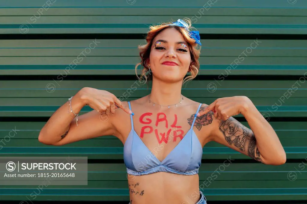 Confident woman showing girl power text on chest in front of green metal wall