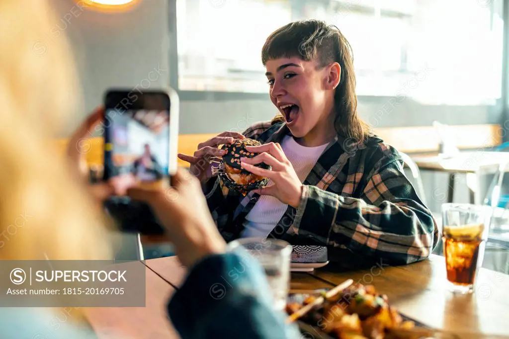 Young woman photographing girlfriend holding burger at restaurant