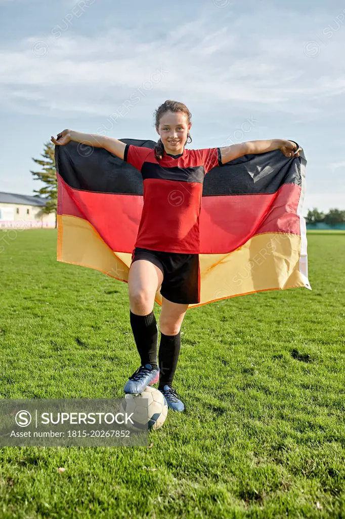 Happy girl standing with soccer ball and holding German flag on field