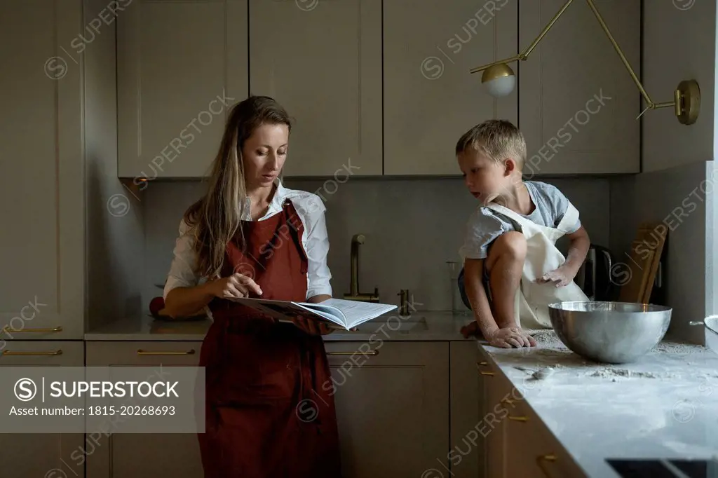 Mother reading recipe book with son sitting on kitchen counter