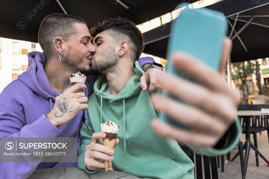 Gay couple kissing and taking selfie over mobile phone at cafe