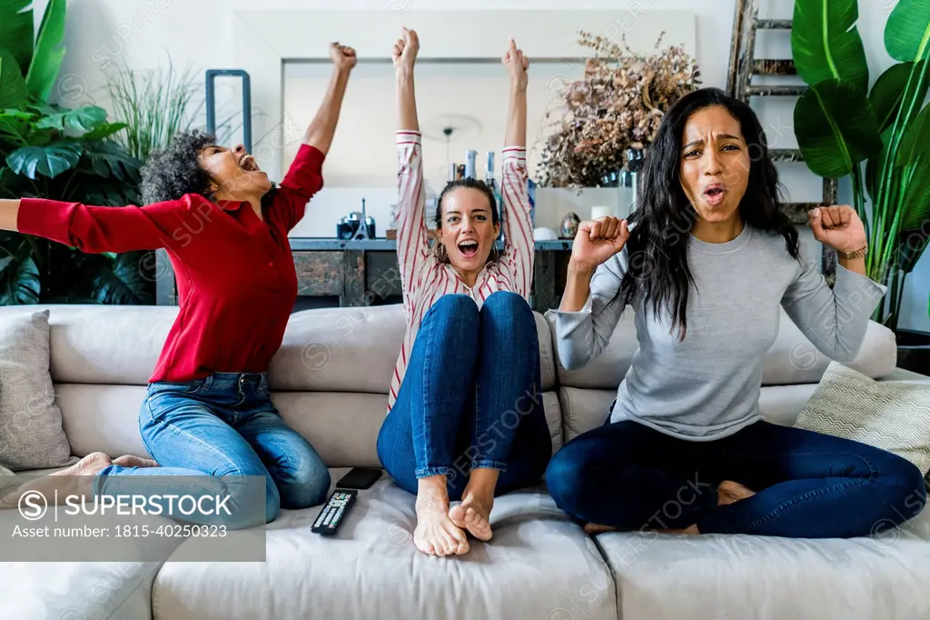 Three excited women on couch at home watching Tv and cheering