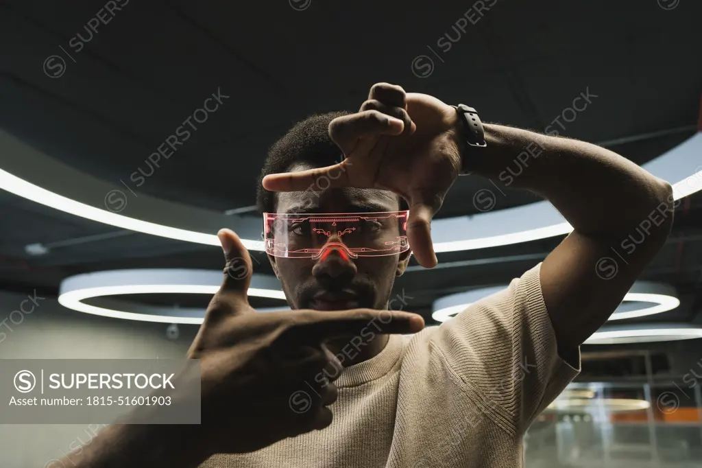 Young man with futuristic cyber glasses under modern ring lamp looking through finger frame