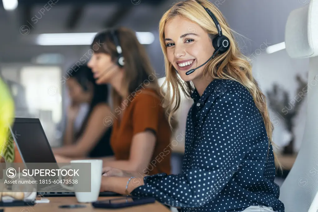 Happy blond businesswoman talking through headset sitting by colleagues at desk in office