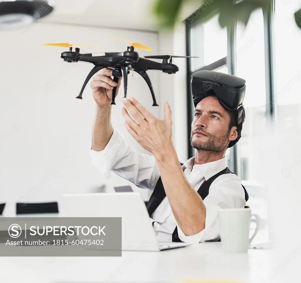 Mature businessman wearing VR glasses and examining drone at desk in office