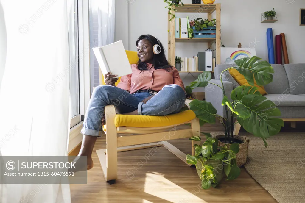 Happy woman listening to music and reading book sitting on chair at home