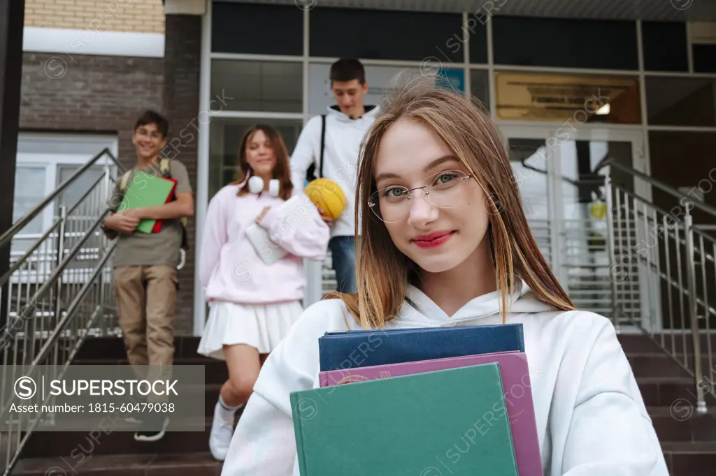 Smiling teenage girl wearing eyeglasses with books standing in front of friends at schoolyard