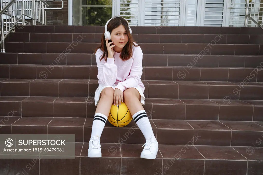 Teenage girl listening to music sitting with ball on steps at schoolyard