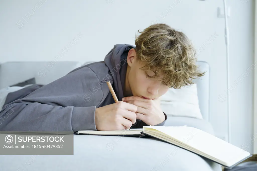 Boy lying on sofa writing in notebook at home