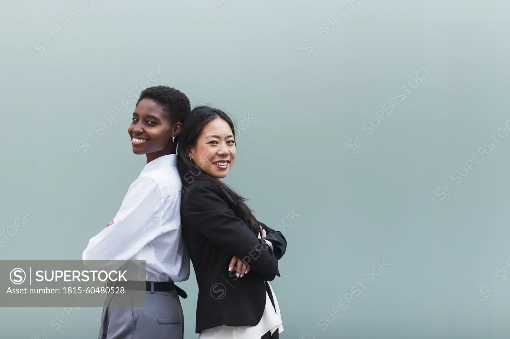 Smiling businesswomen with arms crossed standing back to back in front of blue wall