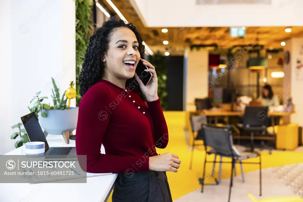 Happy businesswoman talking on smart phone leaning at desk in office