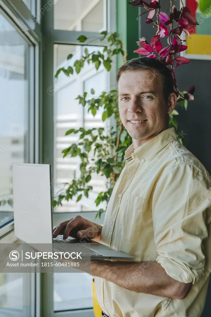 Happy businessman standing with laptop near window in office