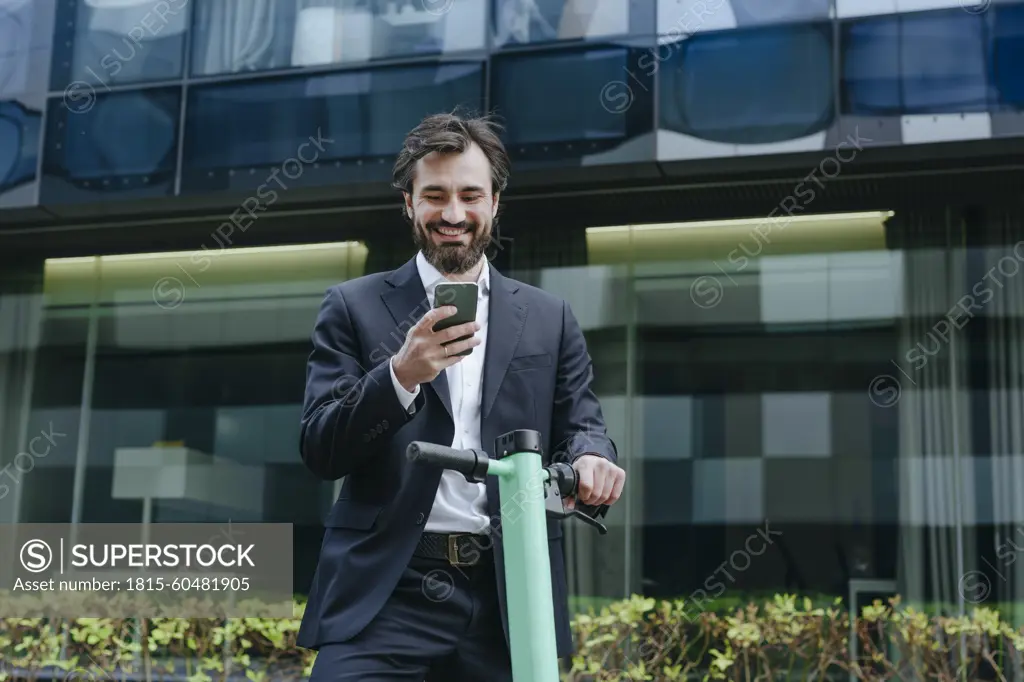 Smiling businessman standing with electric scooter and holding smart phone