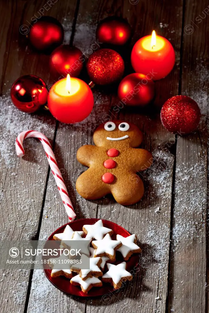 Gingerbread man, sugar cane, plate of cinnamon stars, Christmas baubles and lighted candles on grey wood