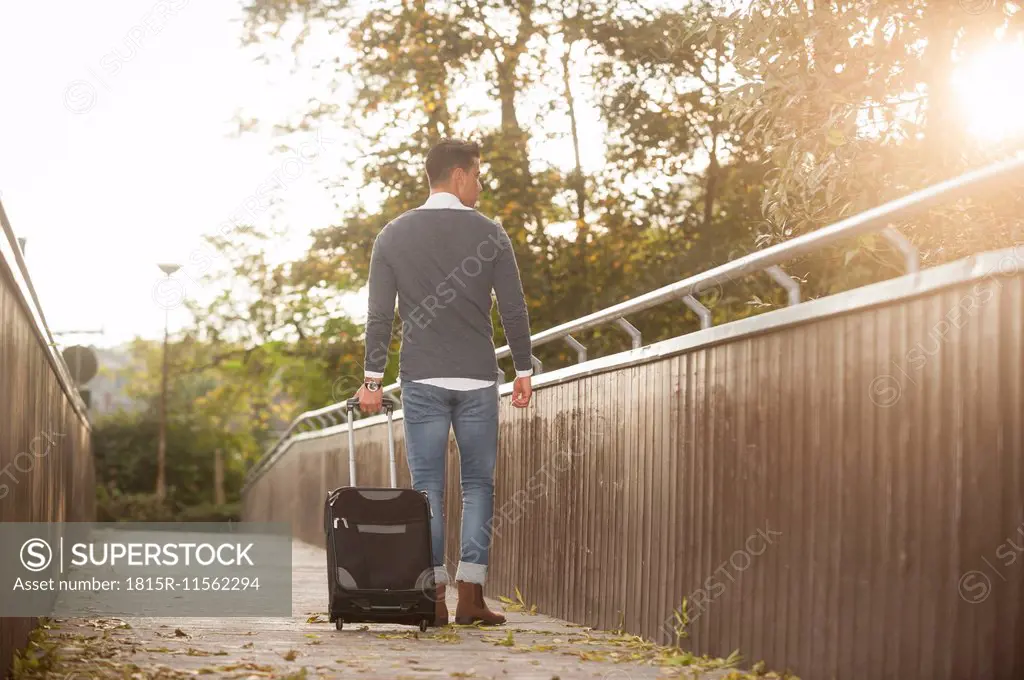 Young man walking with his wheeled luggage on a footbridge at backlight