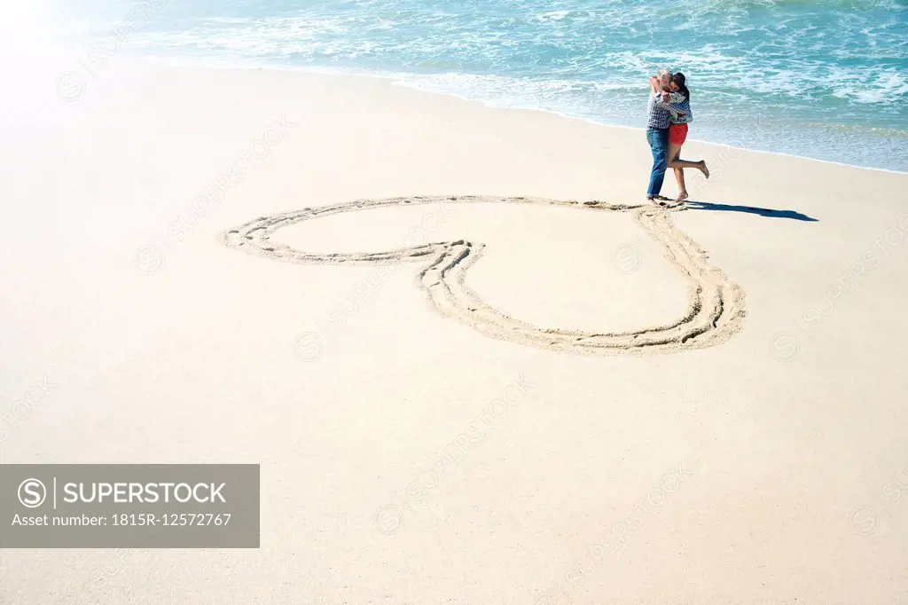 South Africa, couple embracing in front of heart carved in the sand of the beach