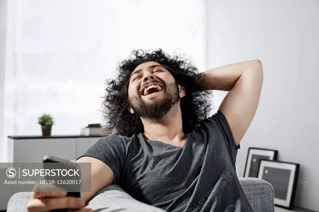 Laughing young man with smartphone sitting on couch at home