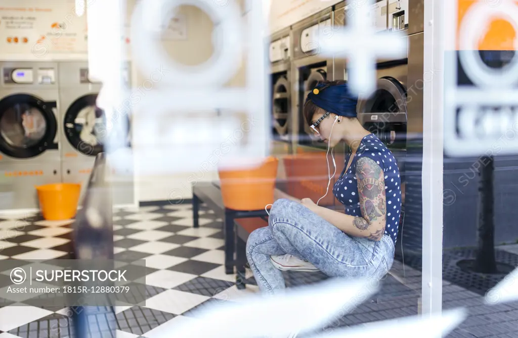 Tattooed young woman hearing music with earphones in a launderette