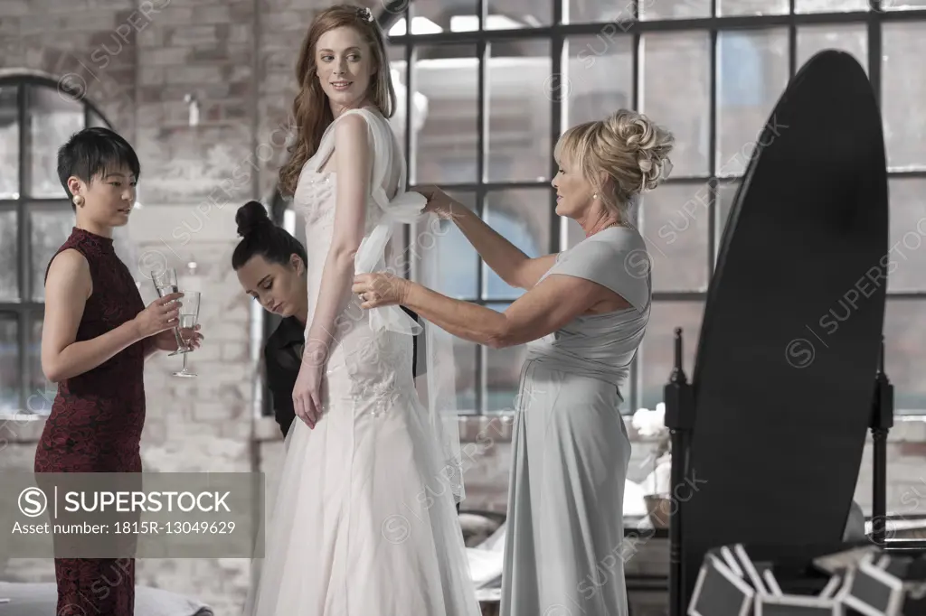 Mother and bridesmaid helping bride putting on wedding dress