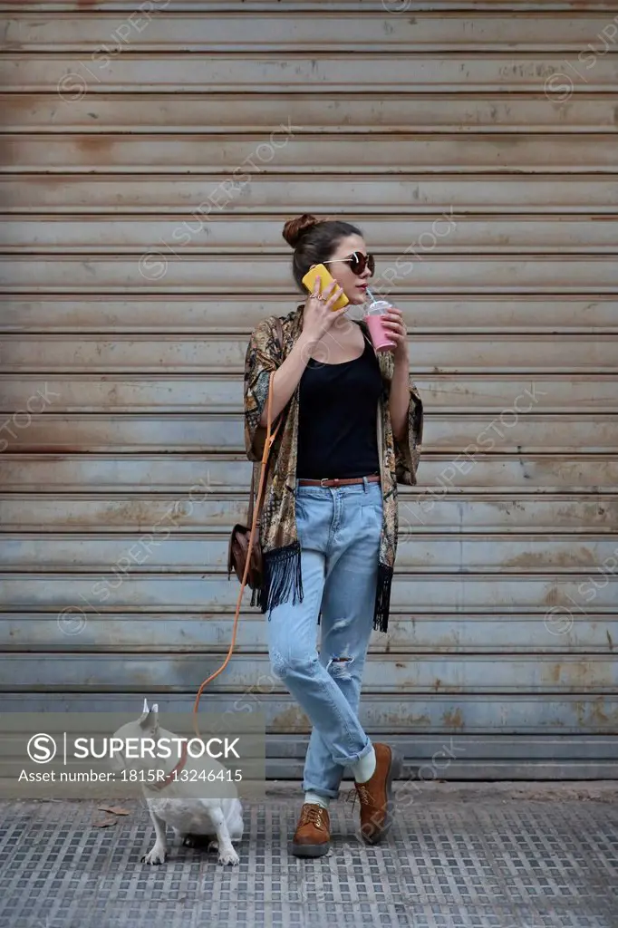 Young woman drinking smoothie and telephoning with smartphone while waiting with her dog on the street