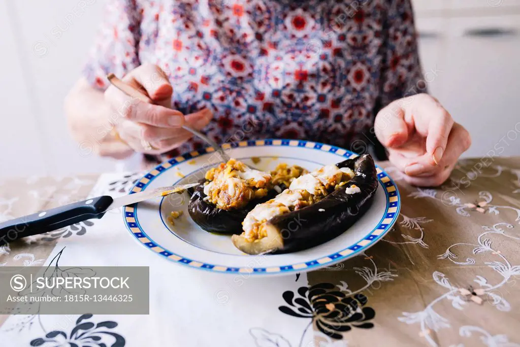 Senior woman eating stuffed eggplant in the kitchen, partial view