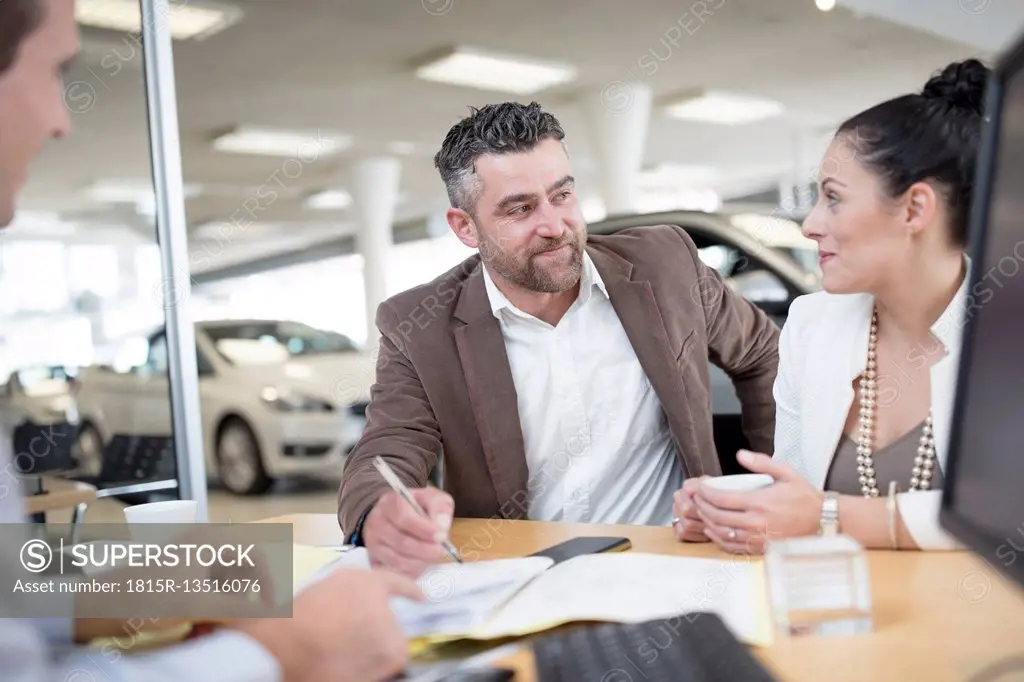 Car dealer and couple sitting at desk with agreement forms in showroom