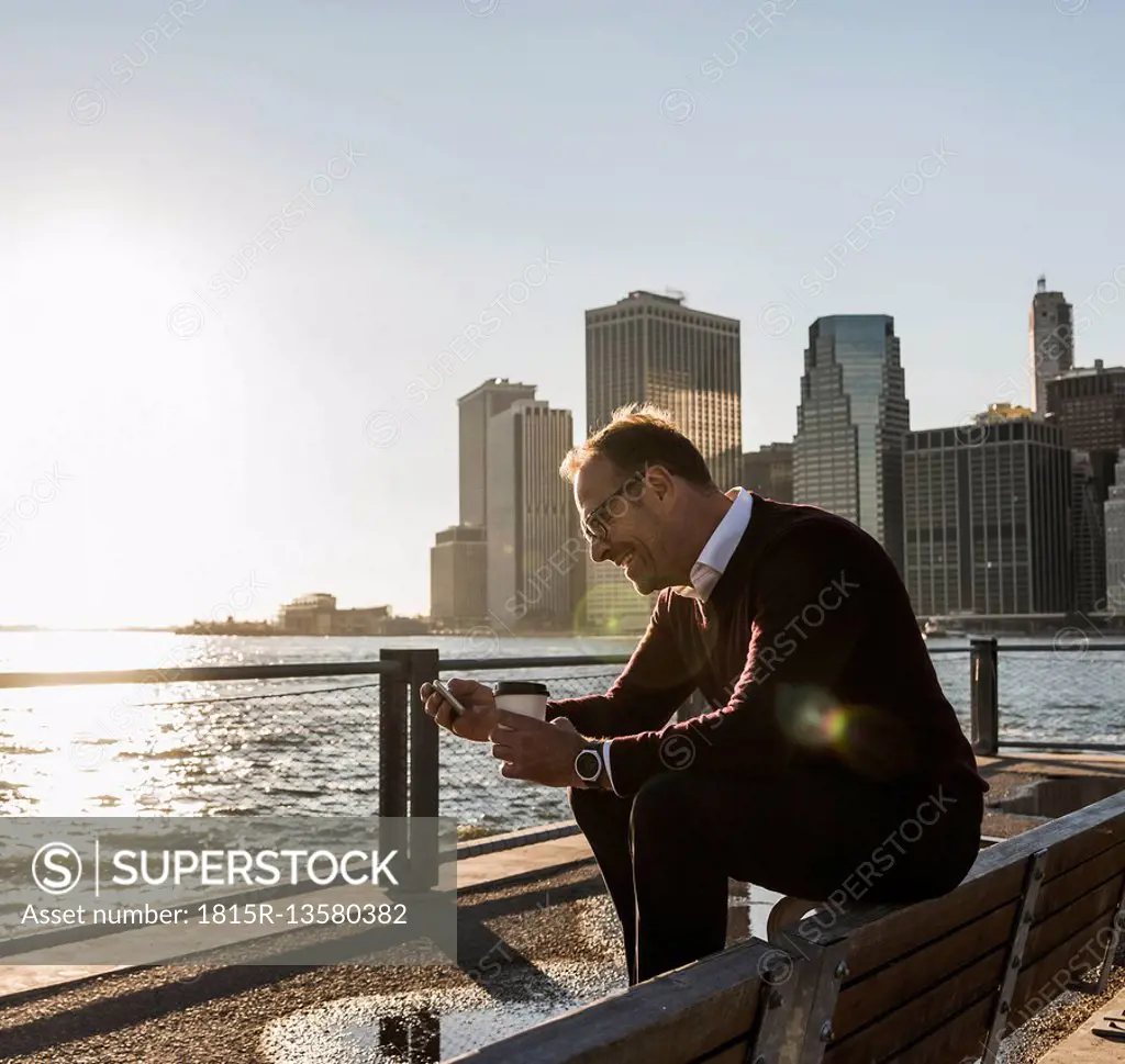 USA, Brooklyn, smiling man with coffee to go sitting on bench looking at smartphone