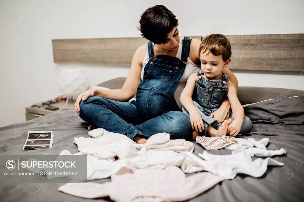 Pregnant woman and her little son sitting together on bed with baby clothing