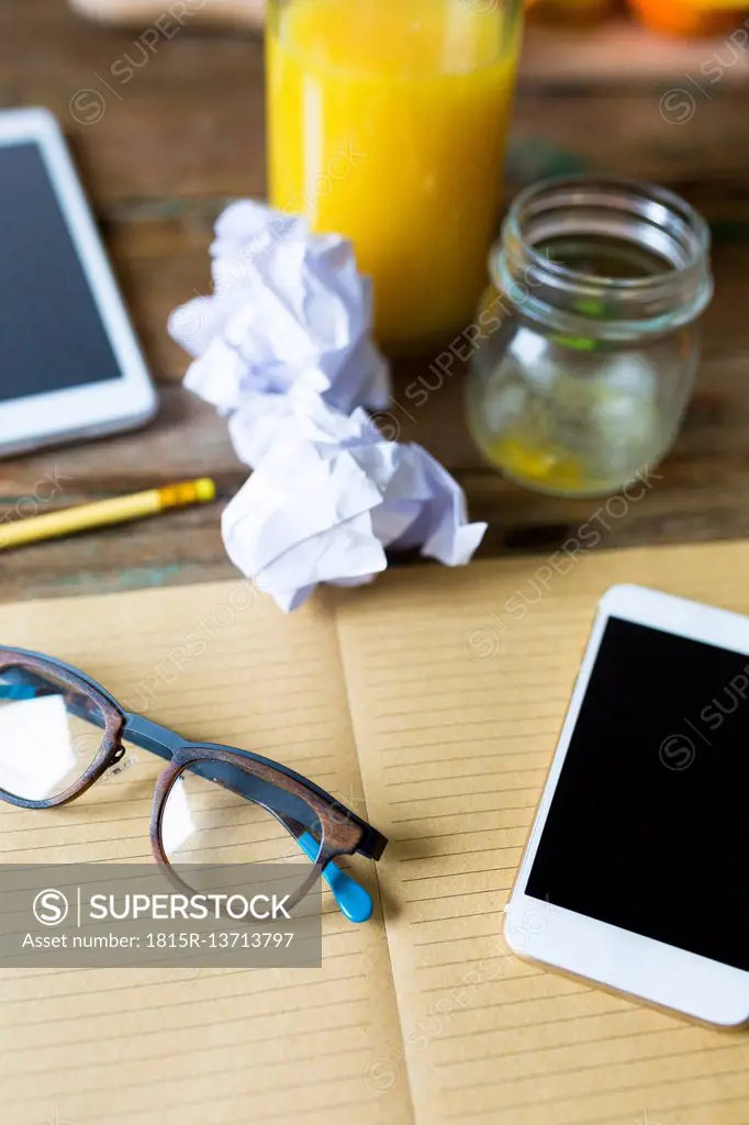 Glass bottle of orange juice, empty glass, notepad, spectacles, tablet, cell phone and crumpled paper